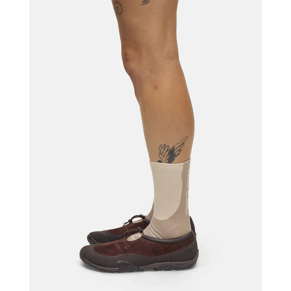 A person stands sideways against a plain white background, wearing Portal’s Lineage All Purpose Socks in Greige and brown shoes. A bird tattoo is visible above one ankle.