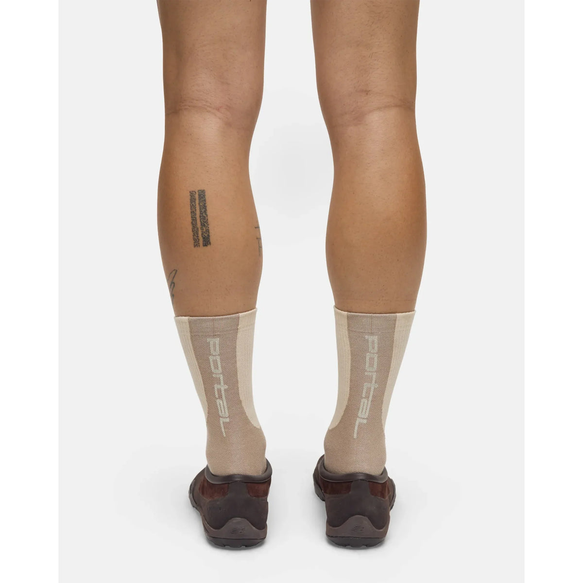 A person with tattooed legs, featuring bar and script designs, stands with their back to the camera on a plain white background, wearing Portal’s Lineage All Purpose Socks in greige—a performance cotton blend offering light compression—paired with brown shoes.