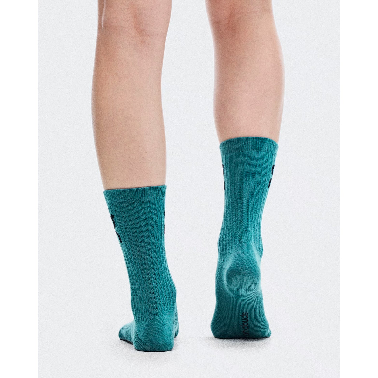 A person stands barefoot except for On’s Logo Socks High in Brook / Storm—a versatile training crew sock—against a plain white background, shown from the back from knees to feet.