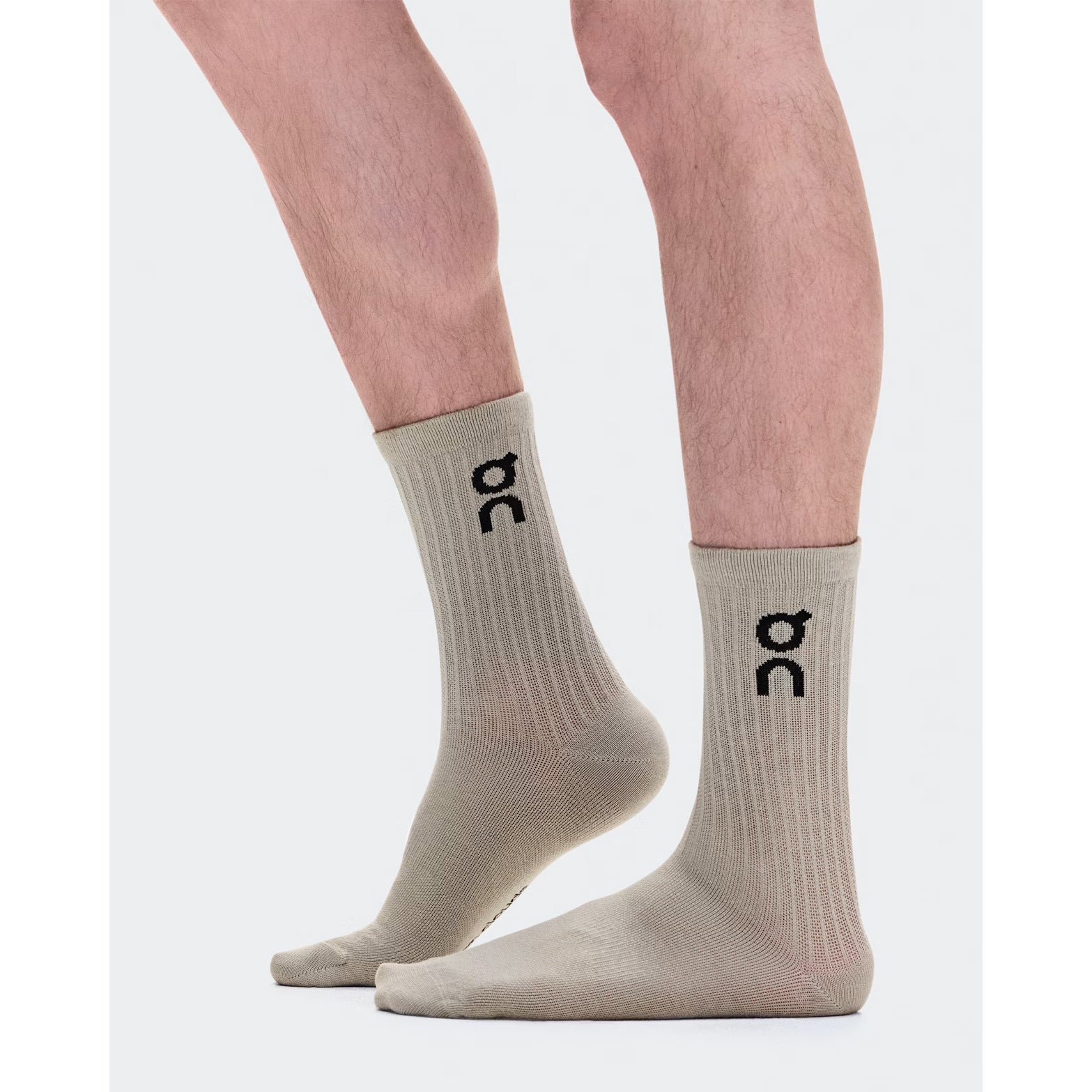 A person is wearing On Logo Socks High in Hare / Brume—a light beige crew sock with a black “On” logo on the sides. These versatile running socks are designed for all-day comfort and arch support, and the person is standing against a plain white background.