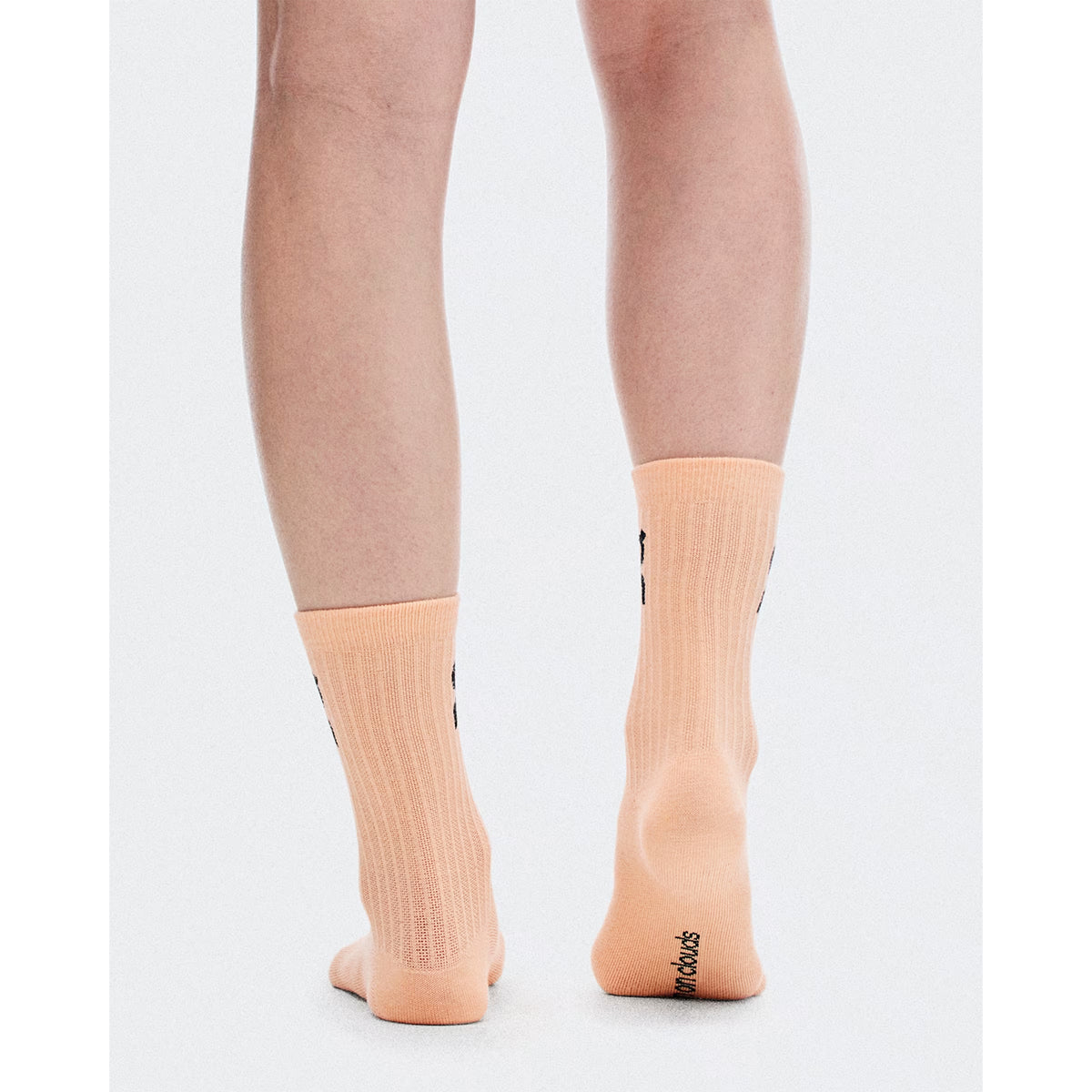 A person stands on a white surface, shown from the calves down, wearing peach-colored Logo Socks High from On in Sherbet / Rhodonite with a ribbed texture and “Mood” text on one heel. These versatile crew socks are designed by On for all-day comfort, seamlessly transitioning from workouts to everyday wear.