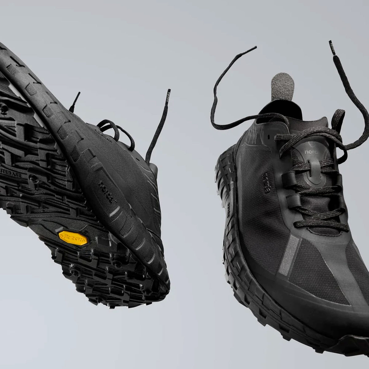 Two black trail running shoes with textured soles are suspended mid-air against a plain light gray background. The shoe on the left displays the yellow Vibram logo on the sole.