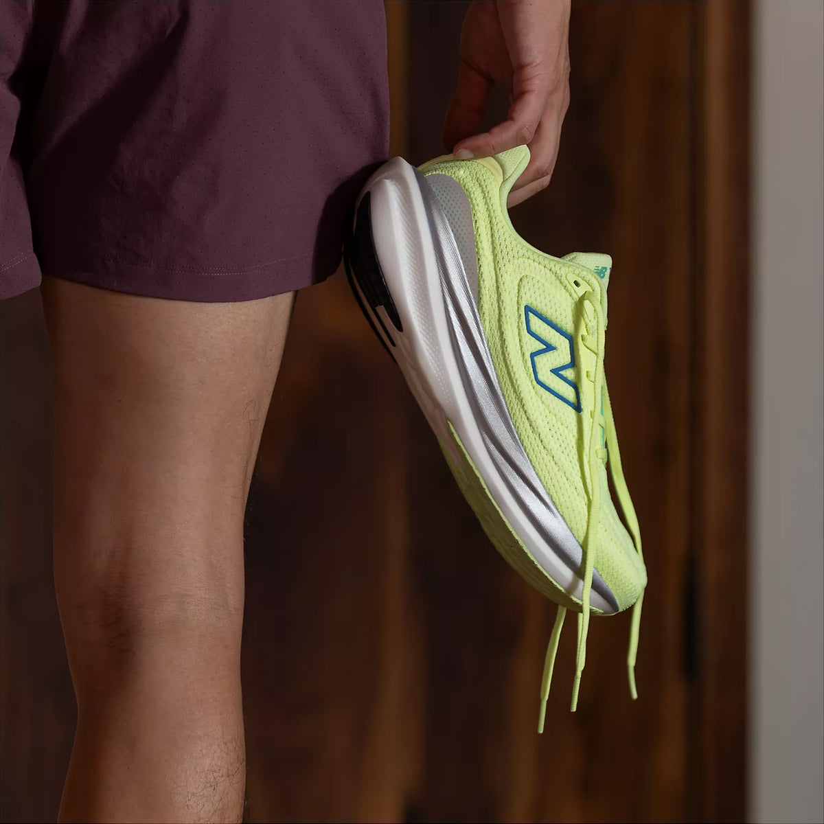 A person wearing maroon shorts holds a yellow New Balance running shoe by the heel, with the shoes blue N logo visible. The background is blurred wood paneling.