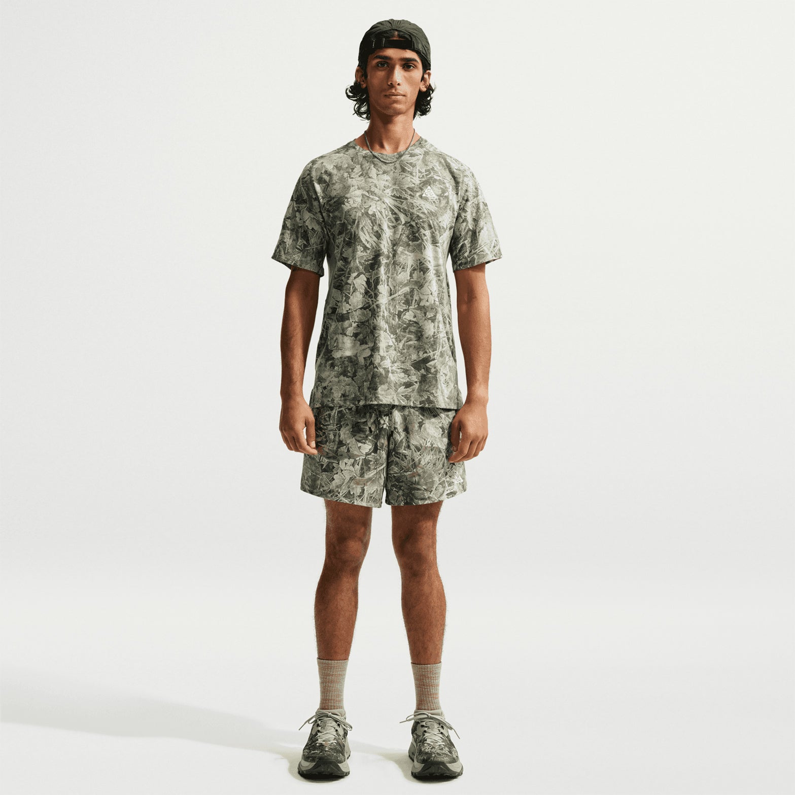 A person stands facing forward, wearing the Nike Men’s ACG Chamo Camo Solar Chase ADV Running Tee in Sequoia/Light Army/Summit White and matching shorts, along with gray socks, sneakers, and a backward black baseball cap. The background is plain white.