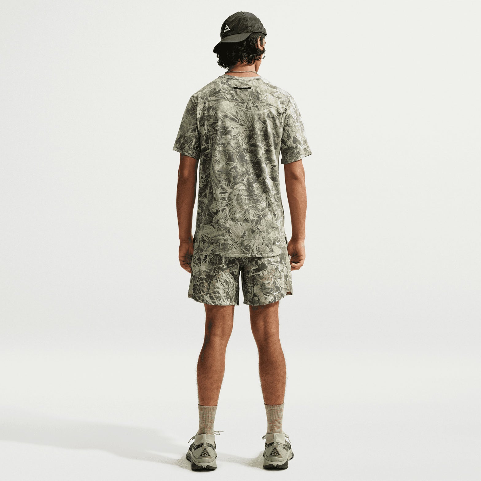 A person stands with their back to the camera, wearing the Nike Men’s ACG Chamo Camo Solar Chase ADV Running Tee in Sequoia / Light Army / Summit White, featuring sweat-wicking Dri-FIT ADV technology. The look is paired with matching gray-patterned running shorts, gray socks, sneakers, and a black cap against a plain white background.