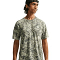A man wearing the Nike Men’s ACG Chamo Camo Solar Chase ADV Running Tee in Sequoia / Light Army / Summit White and a backward black cap looks to the side against a white background.