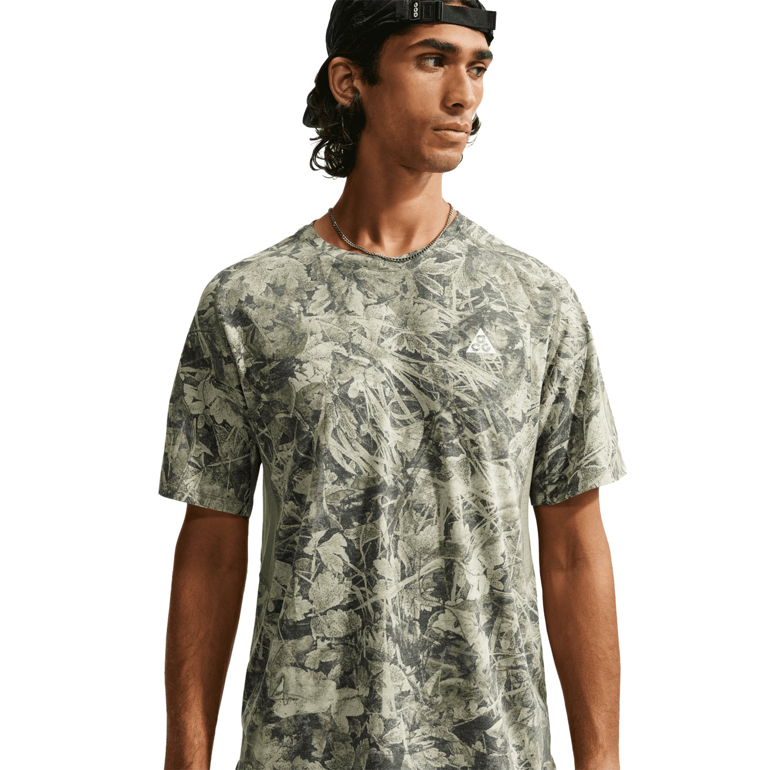 A man wearing the Nike Men’s ACG Chamo Camo Solar Chase ADV Running Tee in Sequoia / Light Army / Summit White and a backward black cap looks to the side against a white background.