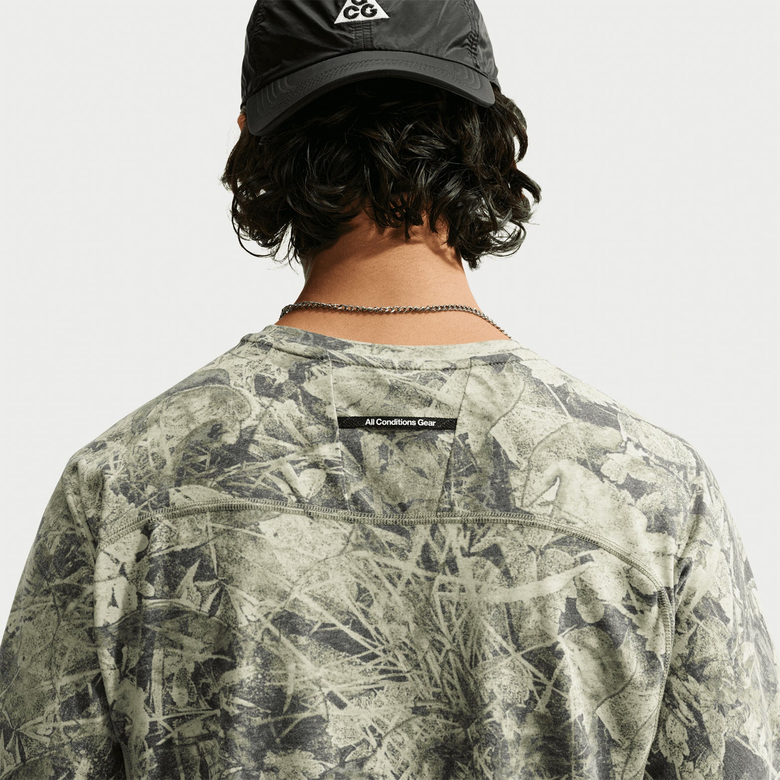 A person wearing a black cap and the Nike Men’s ACG Chamo Camo Solar Chase ADV Running Tee in Sequoia, Light Army, and Summit White with “All Conditions Gear” text on the back, photographed from behind.