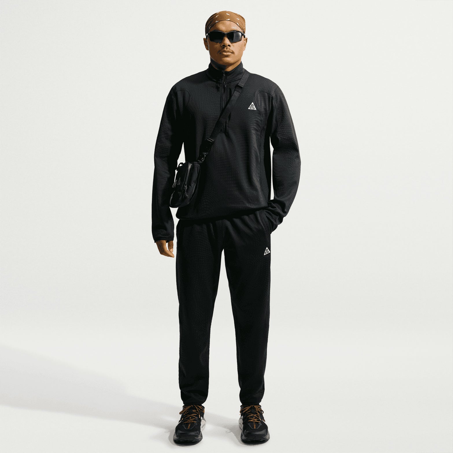 Clad in the Nike Men’s ACG Wolf Grid 1/2 Zip Top in Black / Summit White, along with a crossbody bag, sunglasses, and sneakers, a person stands facing forward against a plain white background—exuding both style and breathable warmth.