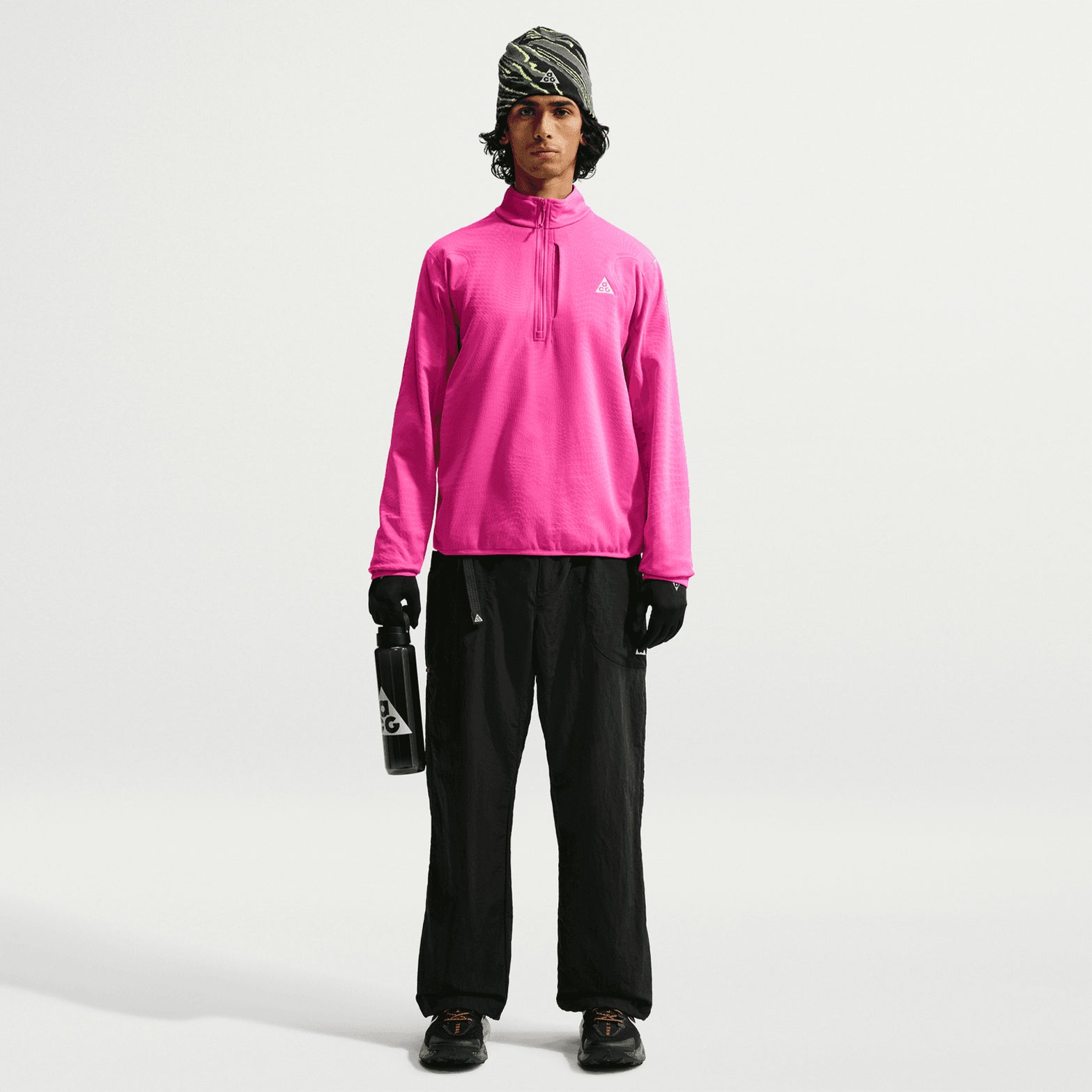 Men's Nike ACG Wolf Grid Therma-FIT 1/2 Zip Top Desert Pink Light Crimson Summit White front view on male trail runner showing half-zip design with thumbholes and vertical zippered chest pocket for hiking and scrambling