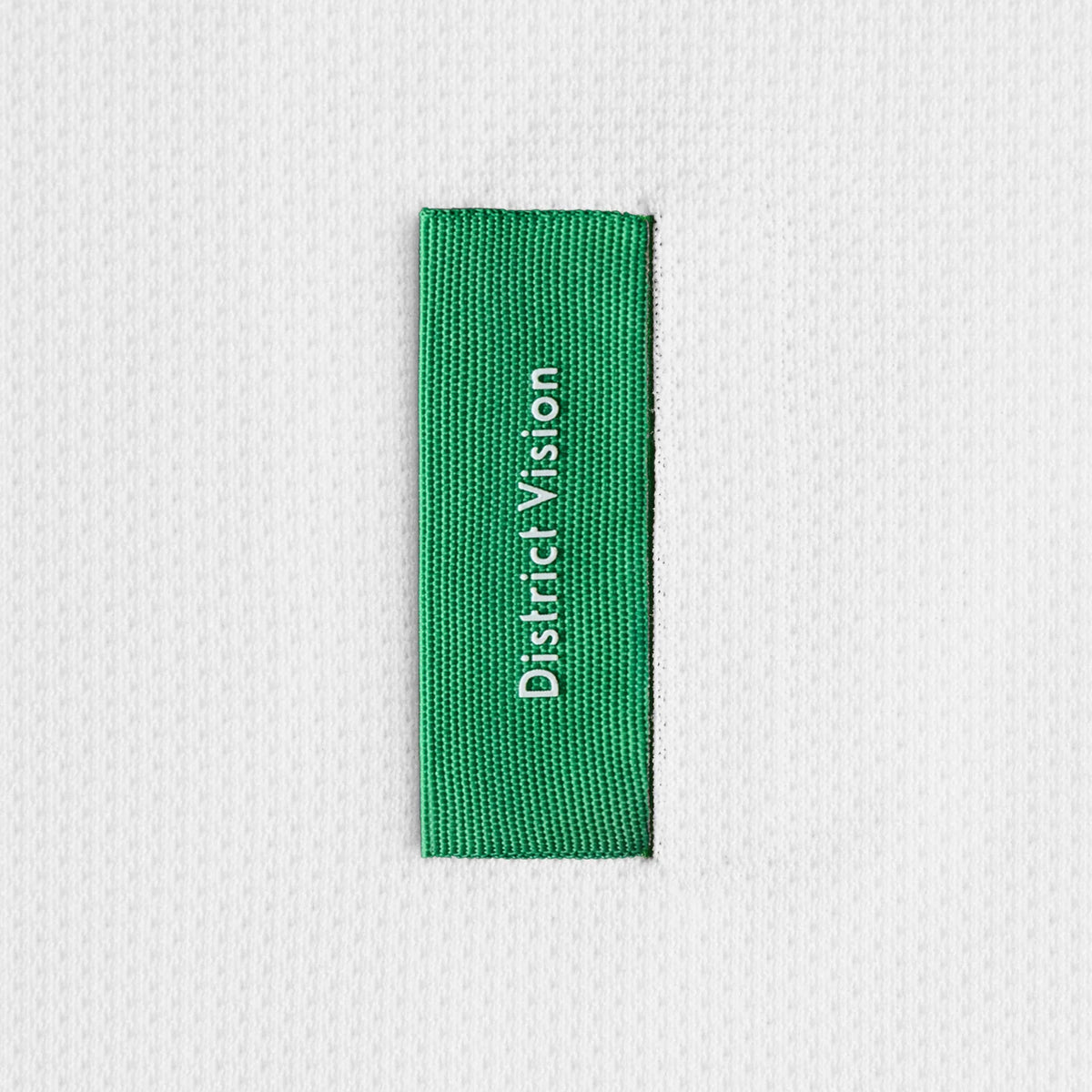 A rectangular green District Vision fabric tag stitched vertically onto the white textured fabric of a Men’s Boxy Training Top Lunar White Flame.