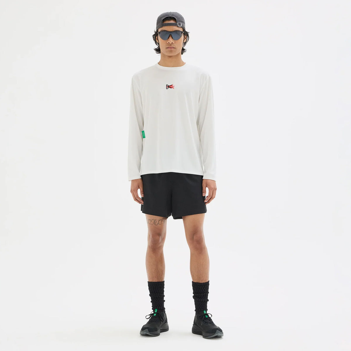 A person wearing a District Vision Men’s Boxy Training Top Lunar White Flame with a small flame logo, black shorts, black athletic socks, sneakers, sunglasses, and a baseball cap.