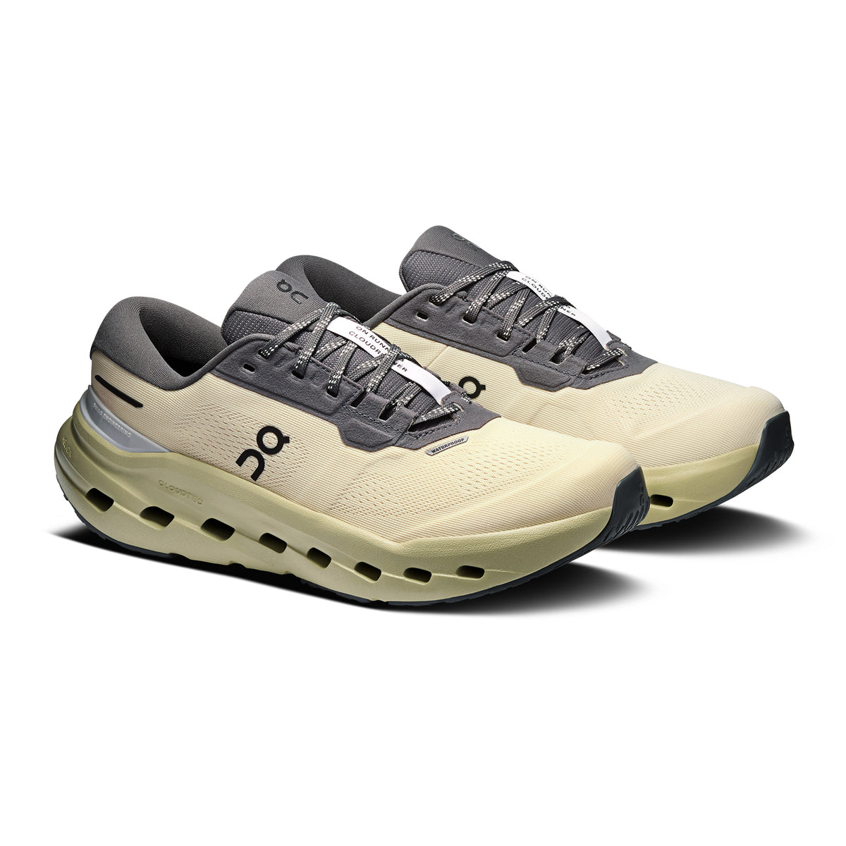A pair of beige and gray athletic running shoes with black accents, thick soles, and visible cushioning. The shoes feature lace-up closures and small logos on the side near the laces.