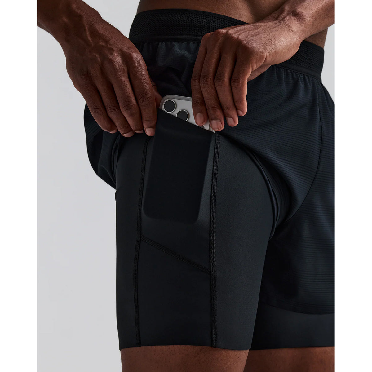 A person in black athletic shorts is placing a smartphone into a side pocket on the shorts. Only the lower torso and upper legs are visible.