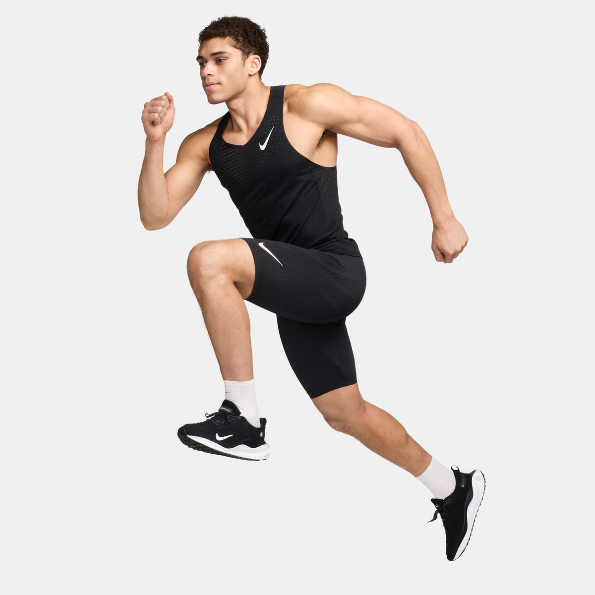 A man in black athletic wear and sneakers is captured in mid-air, appearing to run or jump against a plain white background.