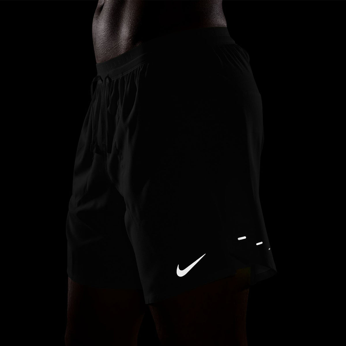 A person wearing black Nike shorts stands against a dark background. Only the lower torso and upper legs are visible, with the white Nike logo prominent on the shorts.