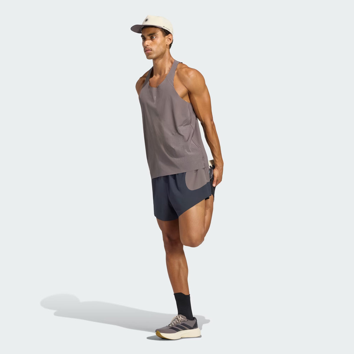 Wearing the Adidas Men’s Hermanos Koumori Running Tank in Charcoal, a man stretches his right leg by holding his ankle behind him and balances on his left leg against a plain light background—perfectly capturing the spirit of performance running.
