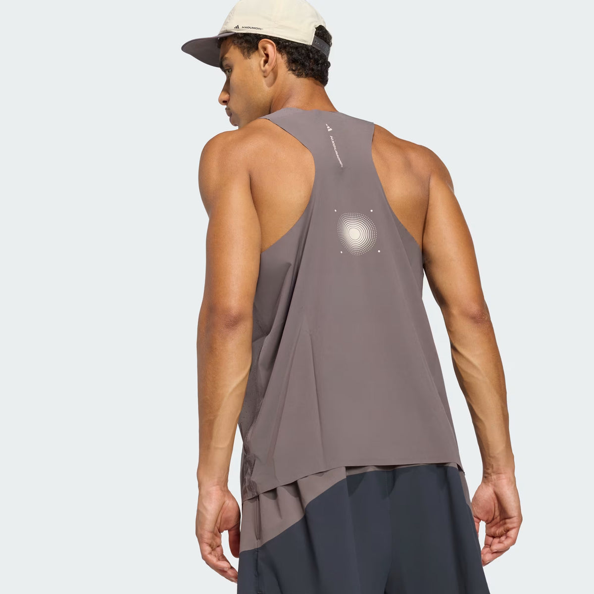A man wearing the Adidas Men’s Hermanos Koumori Running Tank in charcoal, along with black shorts and a light-colored cap, stands facing away to display the back design of his performance running apparel against a plain light background.