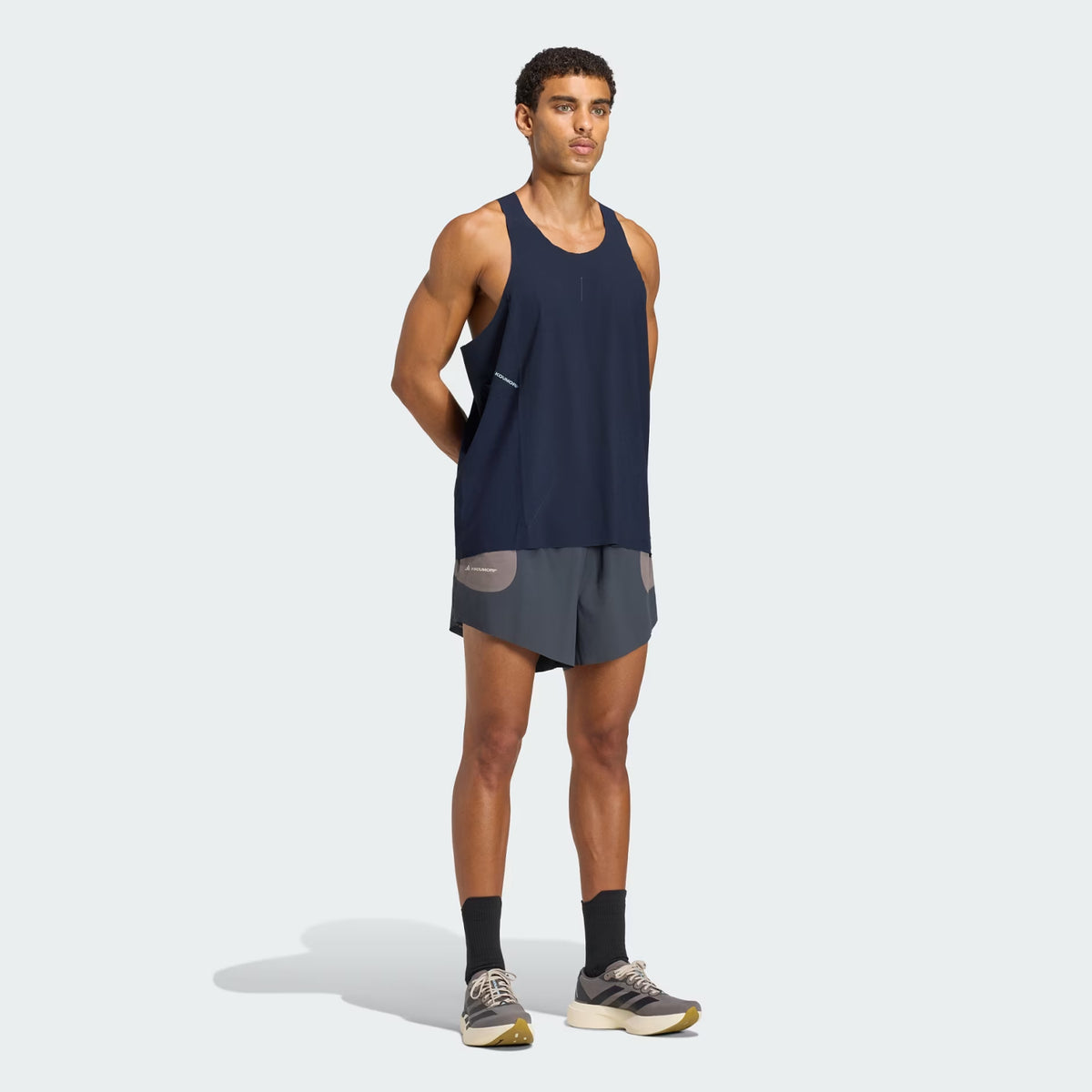 A man wearing a Men’s Hermanos Koumori Running Tank Night Navy, dark grey running shorts, and grey and tan sneakers.