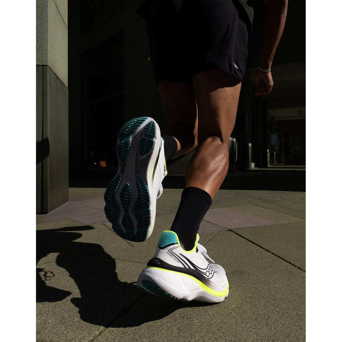 A person wearing black shorts and white running shoes with teal and yellow accents runs on a sunlit city sidewalk, shown from a low rear angle.