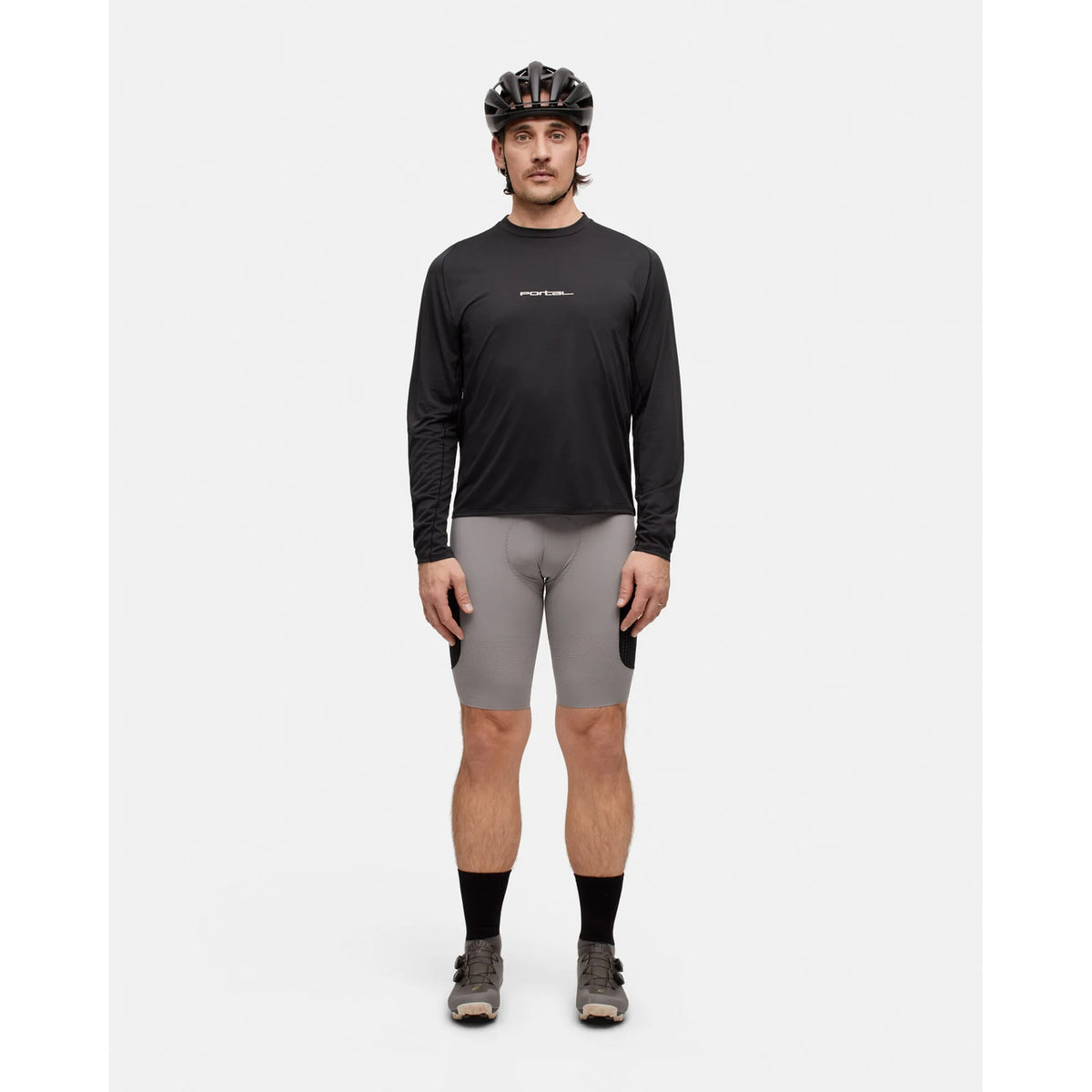 A person wearing a bike helmet, the Portal Men’s Lateral Peak Long Sleeve Top in Black Beauty—a moisture-wicking shirt made from Japanese technical fabric—light gray cycling shorts, black socks, and cycling shoes stands facing forward against a plain white background.