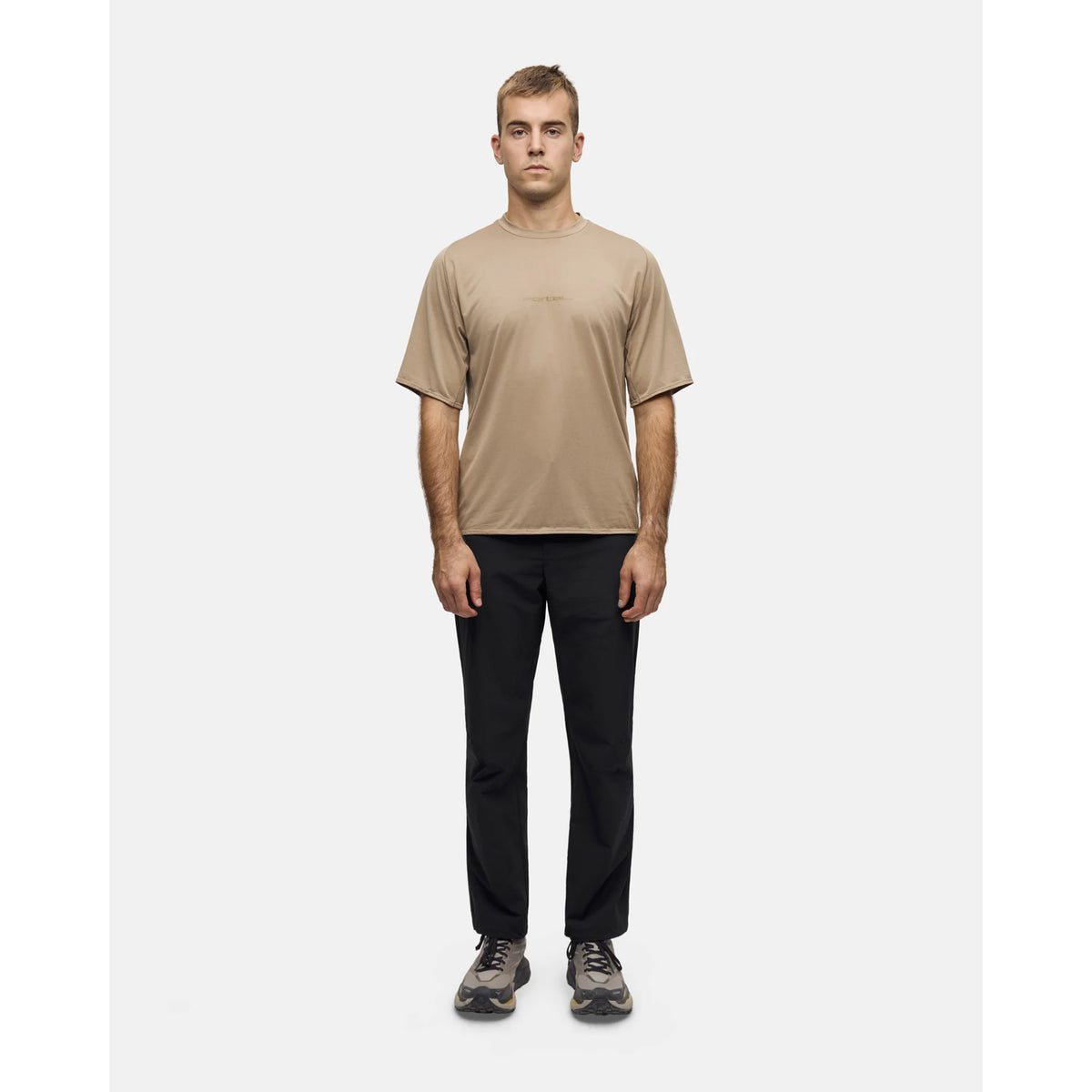 A man wearing a Portal Men’s Lateral Peak Tee Greige tan moisture-wicking technical T-shirt, black pants, and gray athletic shoes.