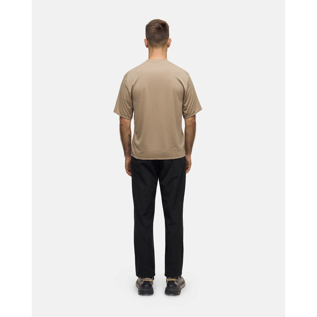 A man seen from behind wearing a short-sleeved, moisture-wicking Portal Men’s Lateral Peak Tee Greige made of recycled polyester, black pants, and athletic sneakers.