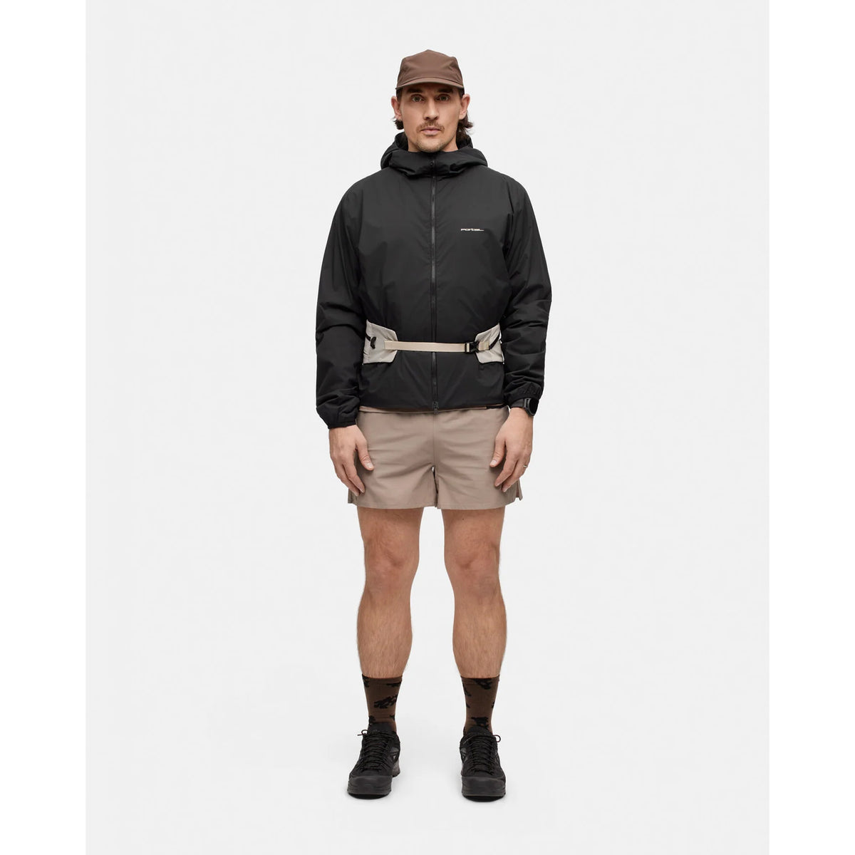 A man wears a black Portal Men’s Level Alpha Jacket Black Beauty with a light-colored utility belt, tan shorts, a brown cap, and dark sneakers.