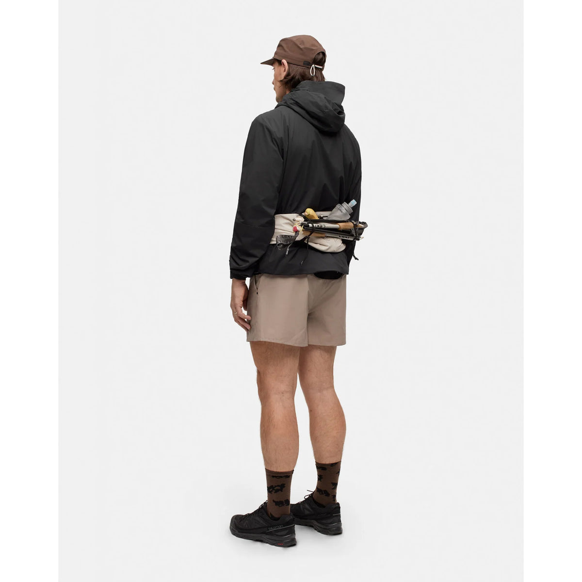 A person seen from behind wears a black Portal Men’s Level Alpha Jacket Black Beauty hooded jacket, tan shorts, and a running belt carrying a water bottle and trekking poles.