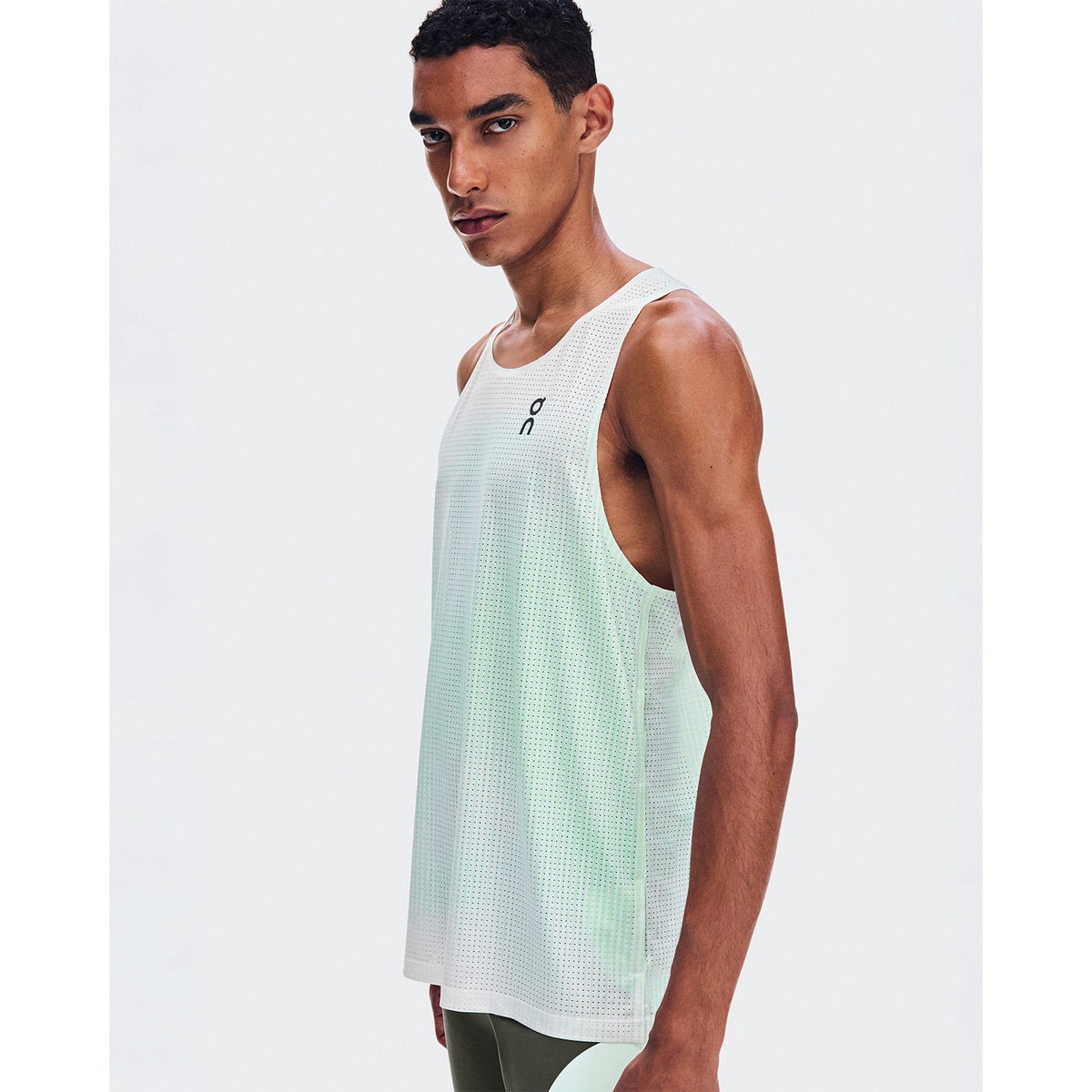A man wearing the On Men’s Race Singlet LN1 Fresco and green pants stands sideways, looking at the camera against a plain white background.