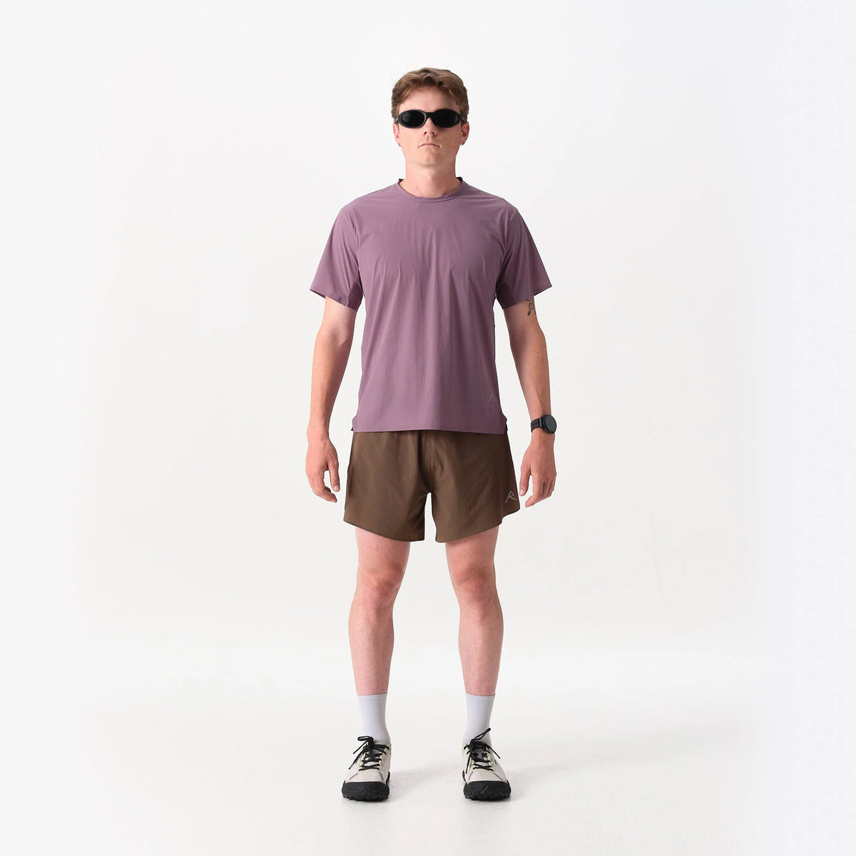 A man wearing a dusty purple Raide Men’s RaceTech Tee Dusk, brown athletic shorts, sunglasses, and sneakers.