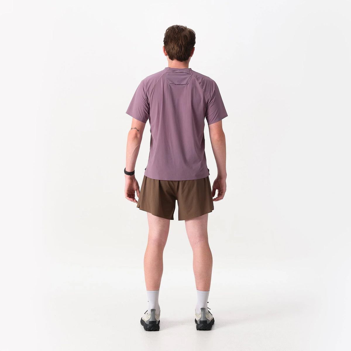 Back view of a person wearing a mauve Raide Men’s RaceTech Tee Dusk, brown running shorts, light-colored crew socks, and trail running shoes.