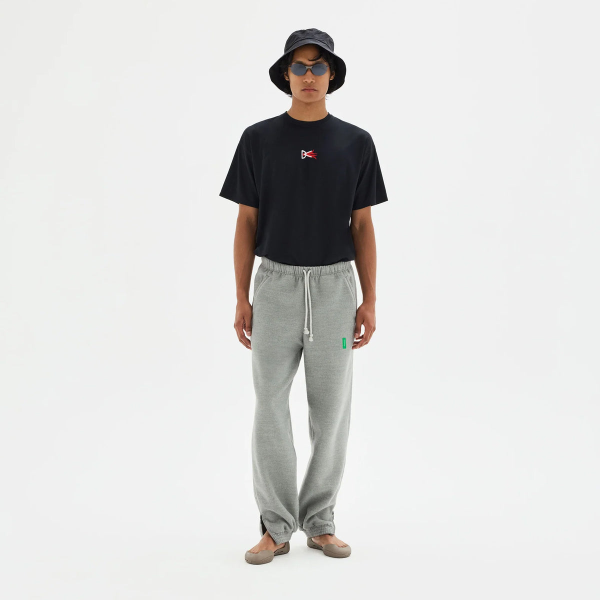 A person wearing a black bucket hat, sunglasses, a District Vision Men’s Short Sleeve Boxy Training Tee Black Flame, and grey sweatpants.