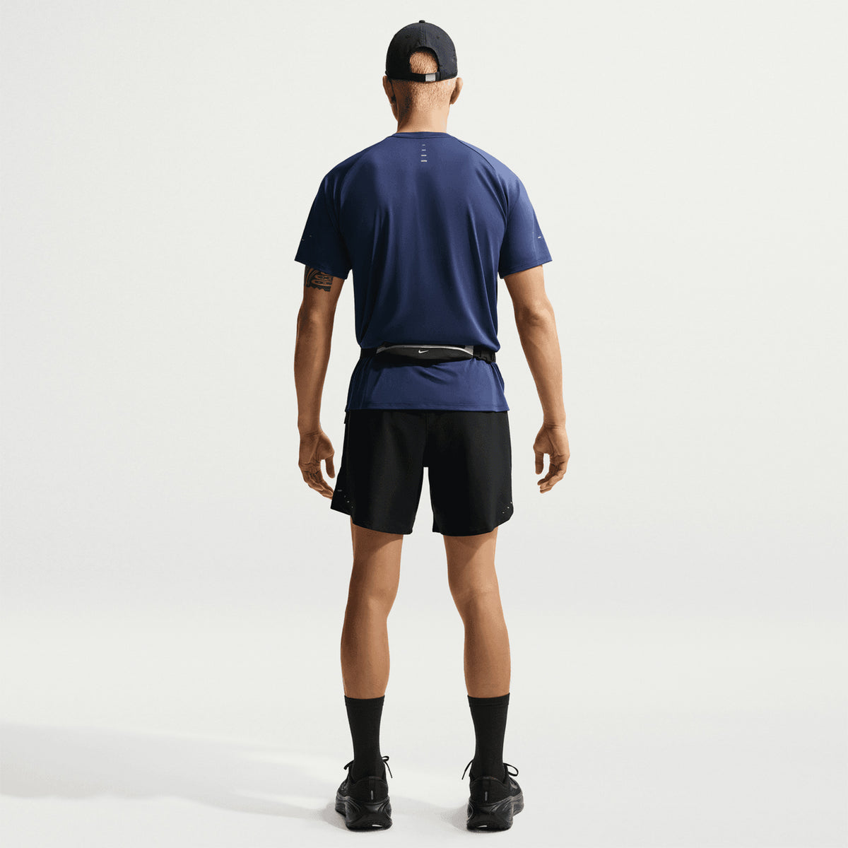 A person stands facing away, wearing a navy blue shirt, Nike Men’s Stride Dri-FIT 7 Inch Running Shorts in black—with a zippered phone pocket—black socks, sneakers, and a black cap. A small waist pouch is strapped around their waist. The background is plain white.