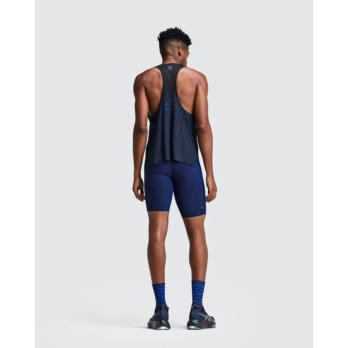 A man viewed from behind wears a black grid-patterned racerback tank top, Bandit Men’s Striped Cadence Half Tights Black / Bauhaus Blue, and patterned crew socks.