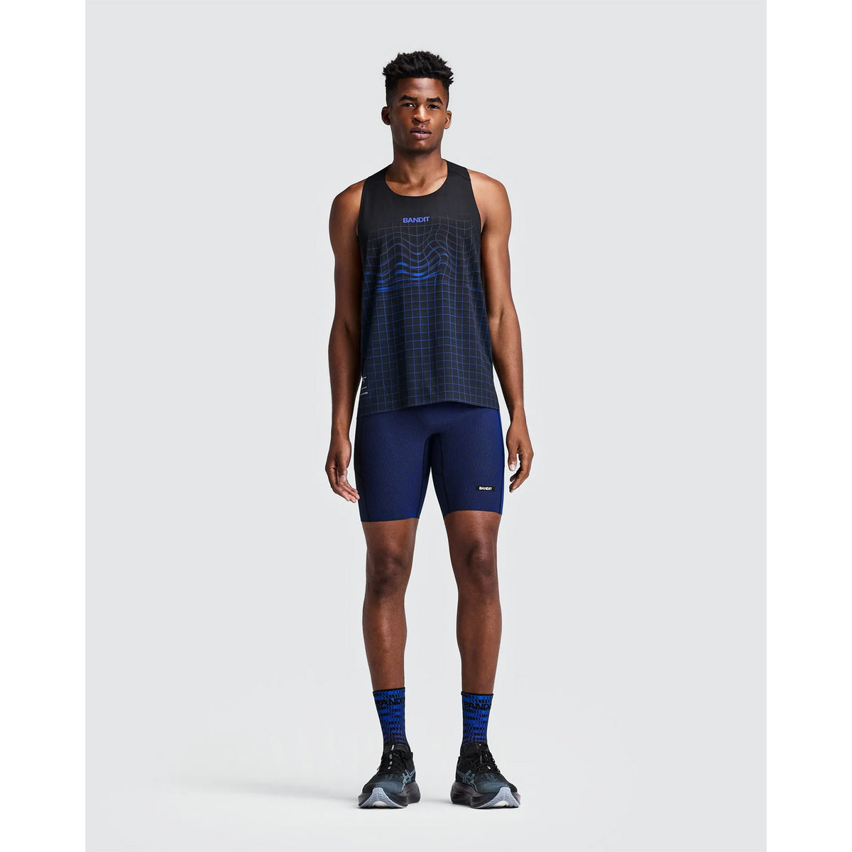 A man wearing a black tank top with blue grid patterns, Bandit Men’s Striped Cadence Half Tights Black / Bauhaus Blue, and matching blue-patterned socks with black running shoes.