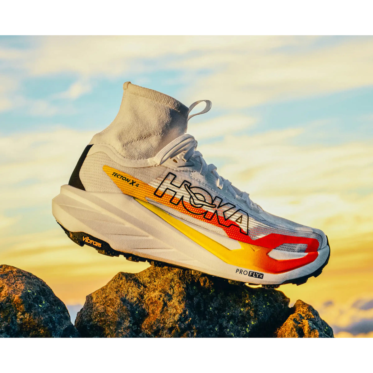 A white HOKA running shoe with red and yellow accents sits on rocks at sunset, displaying the Vibram sole and PROFLY X label against a colorful sky.
