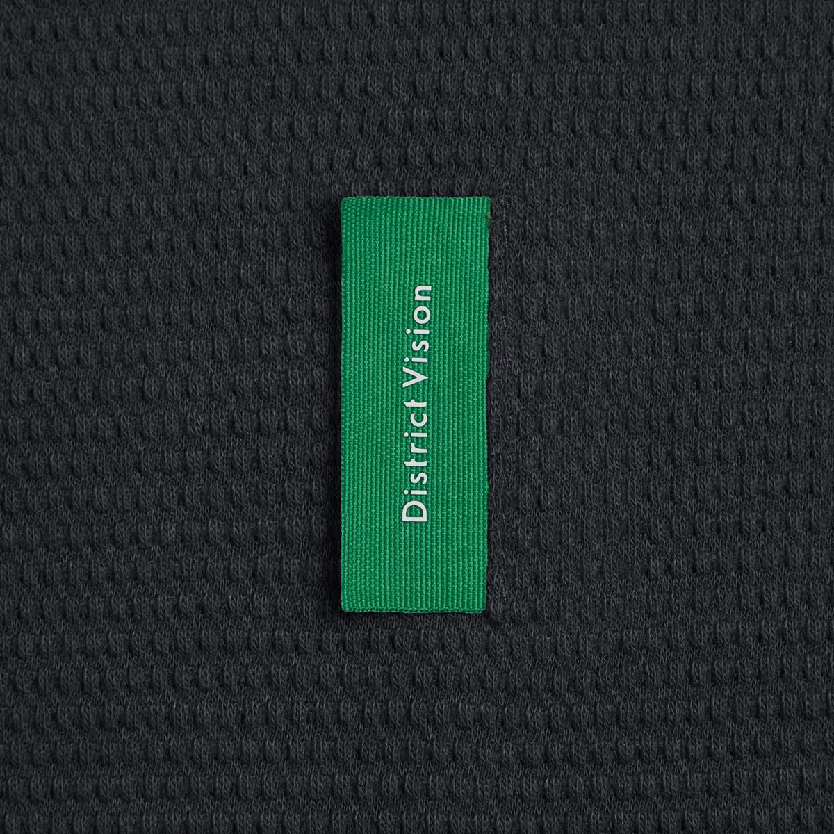 Green vertical rectangular label with white District Vision text sewn onto the black, waffle-textured knit fabric of a Men’s Waffle Top Black.