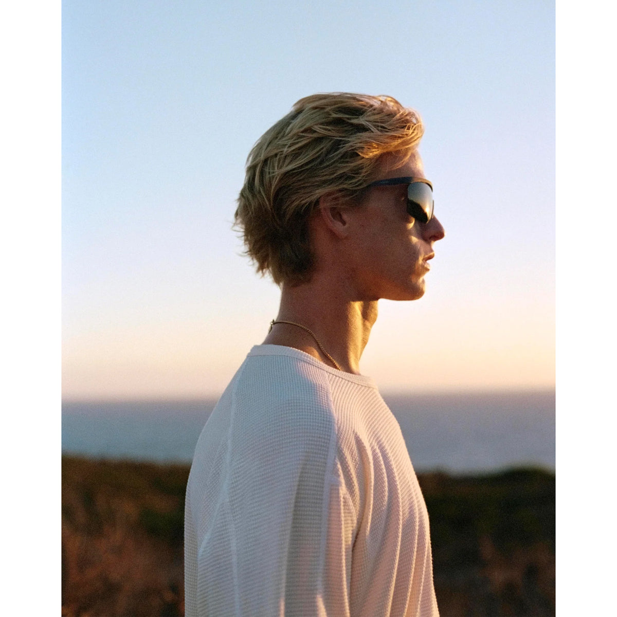A man wearing a white District Vision Men’s Waffle Top and sunglasses looks out over the ocean at sunset.