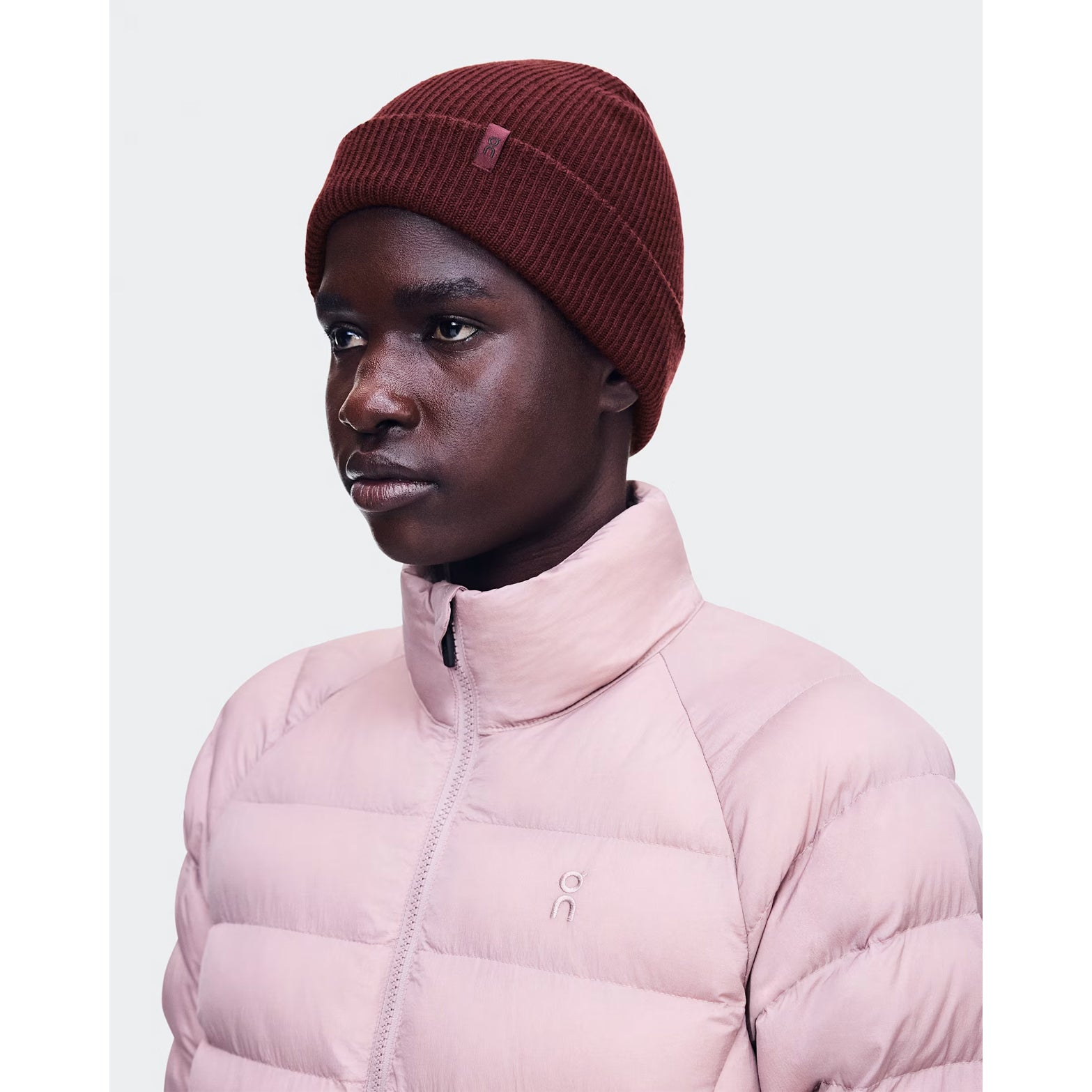 Wearing the On Merino Beanie Maroon and a light pink puffer jacket, a person stands against a plain light background, looking slightly to the side—an ideal combination for comfortable temperature regulation.