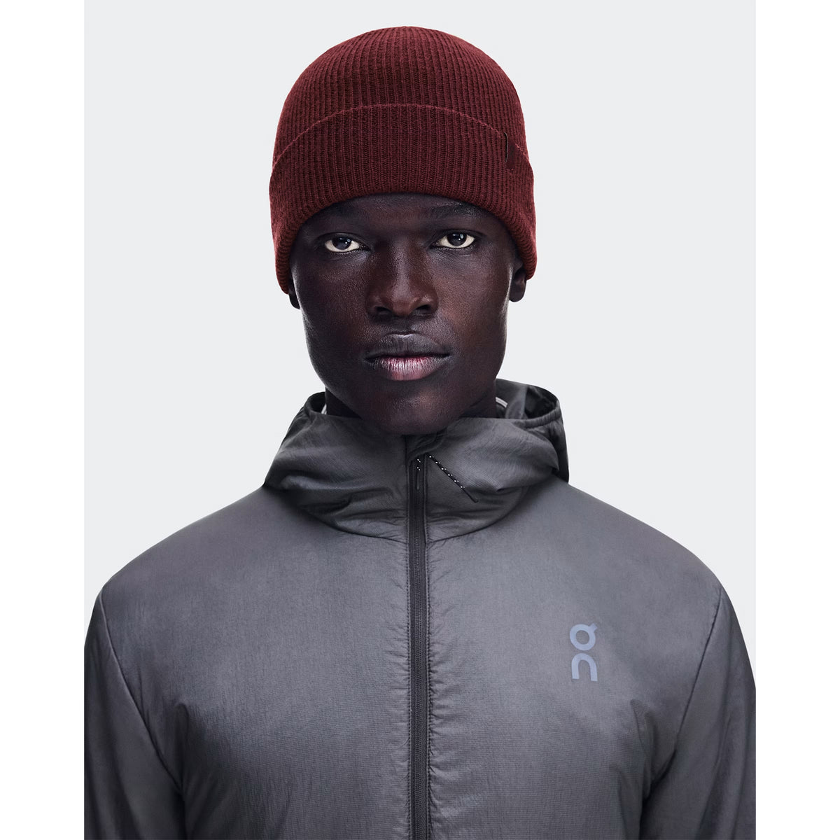 A person wearing the On Merino Beanie in Maroon and a gray zip-up jacket with an On logo, looking straight ahead against a plain light background.