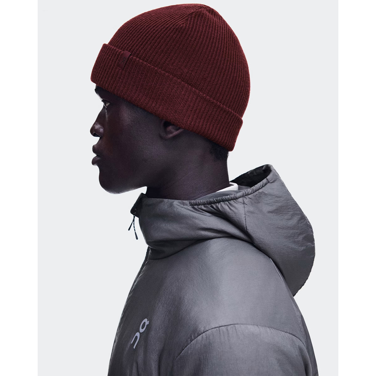 A person in profile wears the On Merino Beanie Maroon and a gray hooded jacket, looking to the left against a plain light background, effortlessly balancing warmth and temperature regulation.