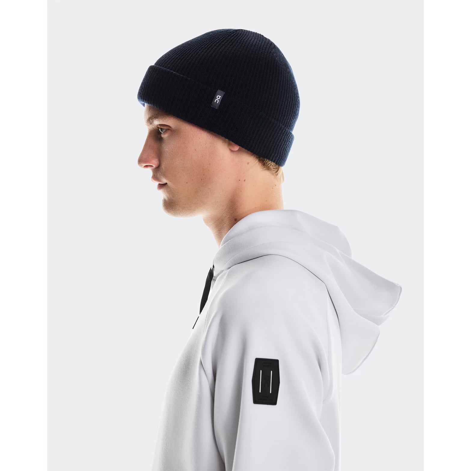 A person in the On Merino Beanie Midnight and a white hoodie with a black rectangular sleeve patch faces left against a plain light background.