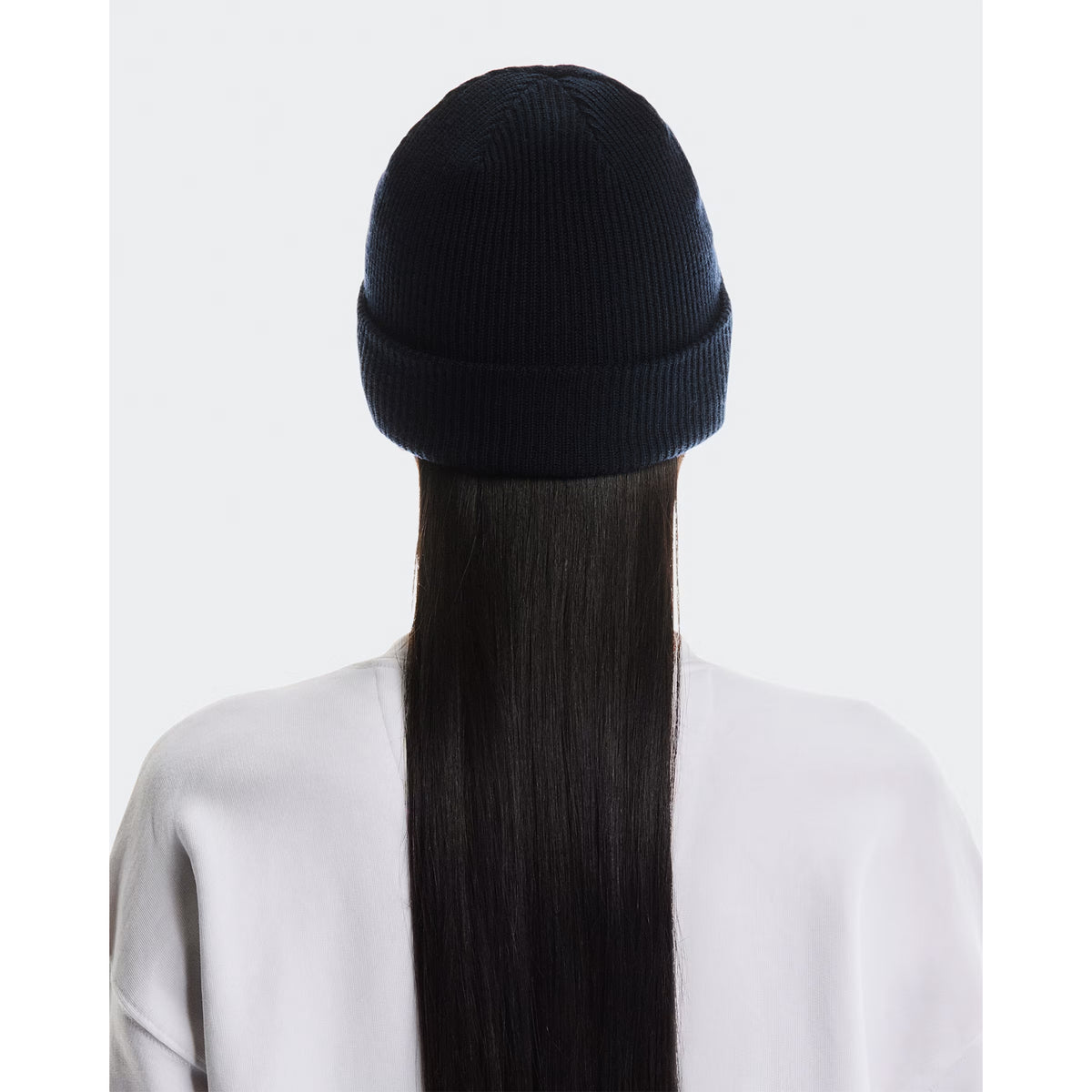 A person with long, straight black hair is shown from the back against a plain light gray background, wearing the On Merino Beanie Midnight and a white shirt, highlighting both style and temperature regulation.
