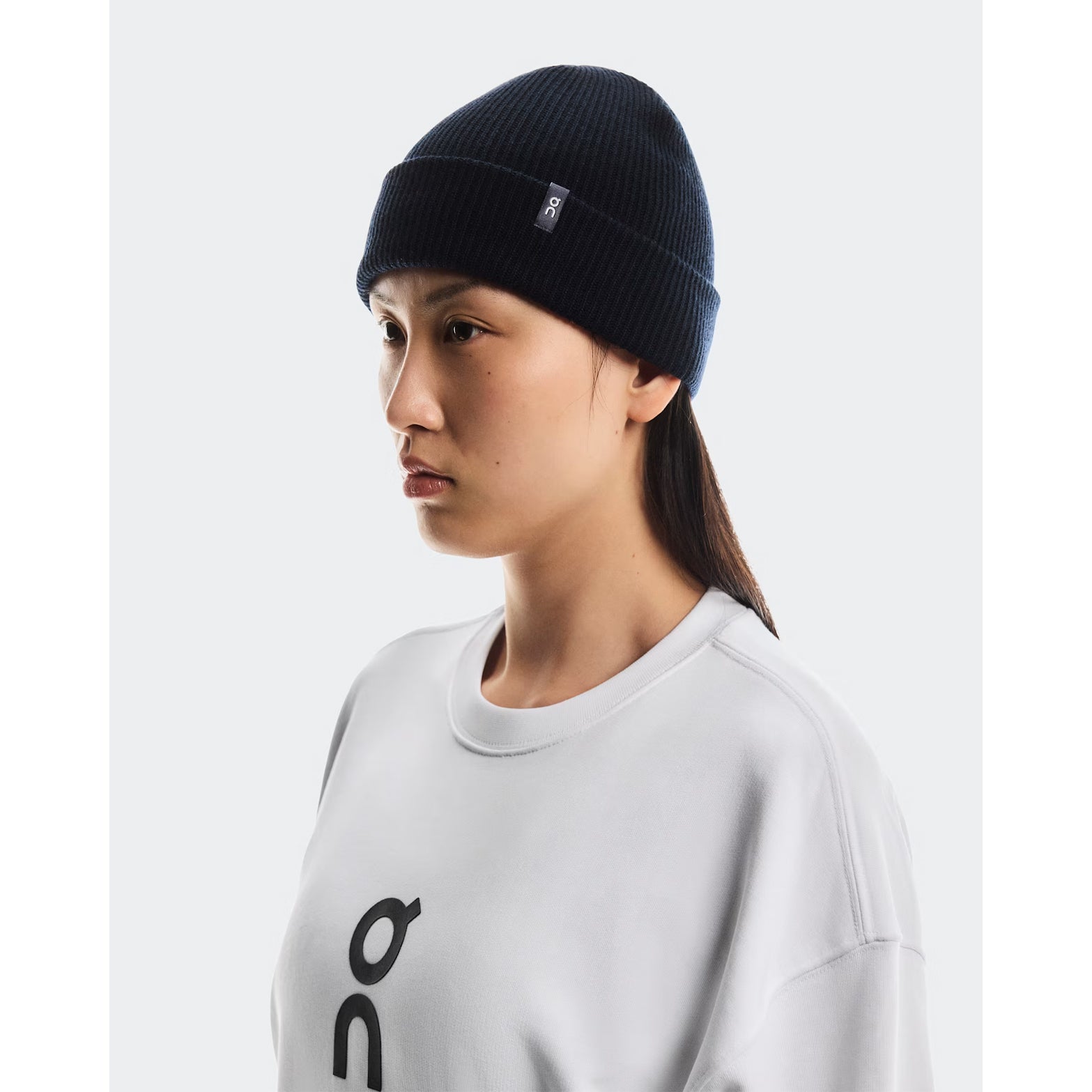 A person is shown in profile, facing left, wearing the On Merino Beanie Midnight and a light gray sweatshirt with a black logo, standing against a plain white background.