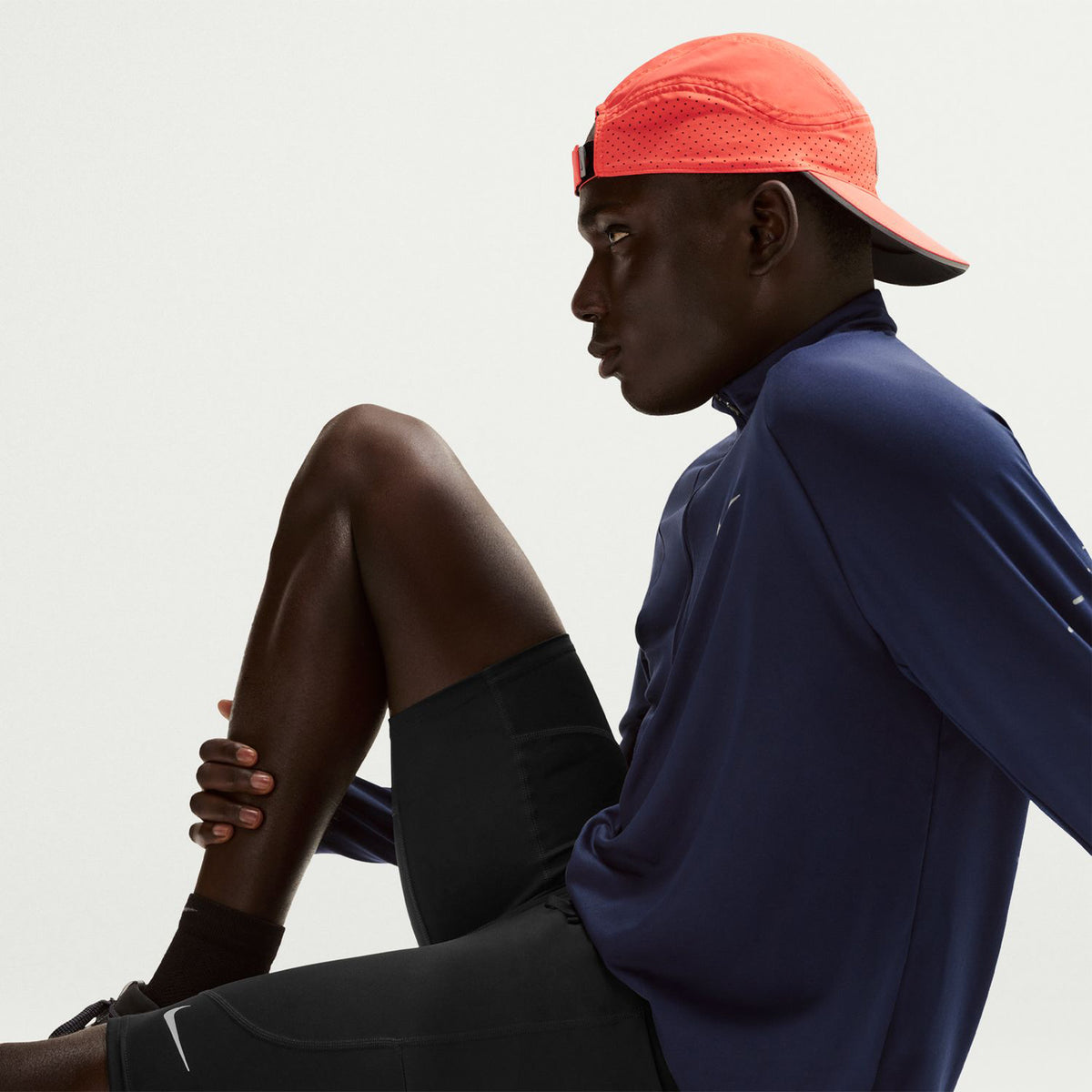 A person wearing a coral cap backward, a navy long-sleeve shirt, and black shorts sits with one knee raised, looking to the side against a plain light background.