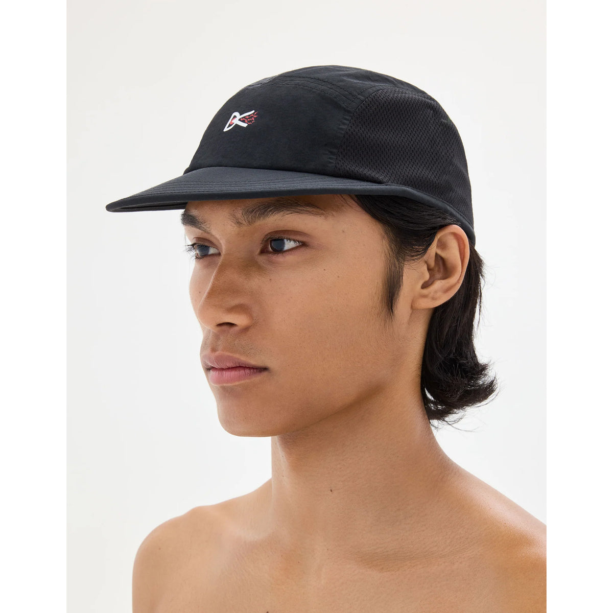 A person wearing a black District Vision Nylon Paneled Mesh Hat, a water-resistant five-panel running hat featuring a small red and white logo on the front.