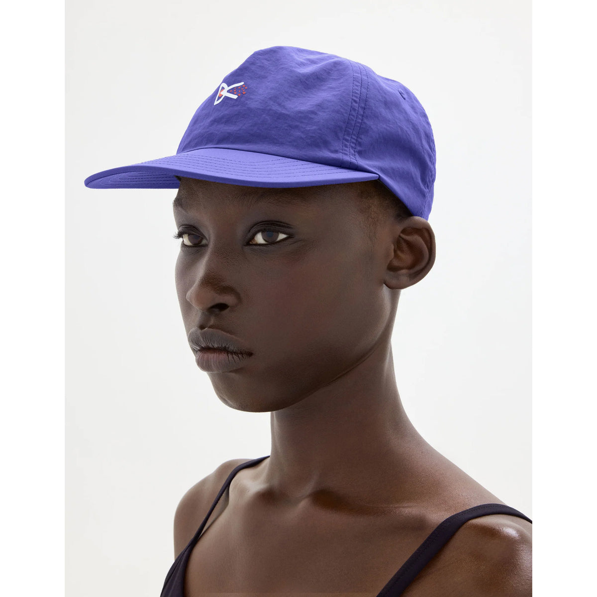 A person wears a Paneled Nylon Cap Symbol Vibrant Lilac, a vibrant lilac, ultralight nylon cap featuring a small white and red logo and an adjustable strapback closure.