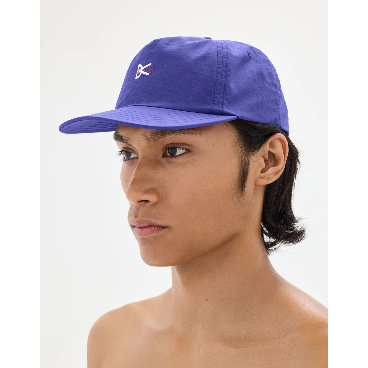 A person wearing a District Vision Paneled Nylon Cap Symbol Vibrant Lilac, featuring a small white and red logo on the front panel and an adjustable strapback closure.