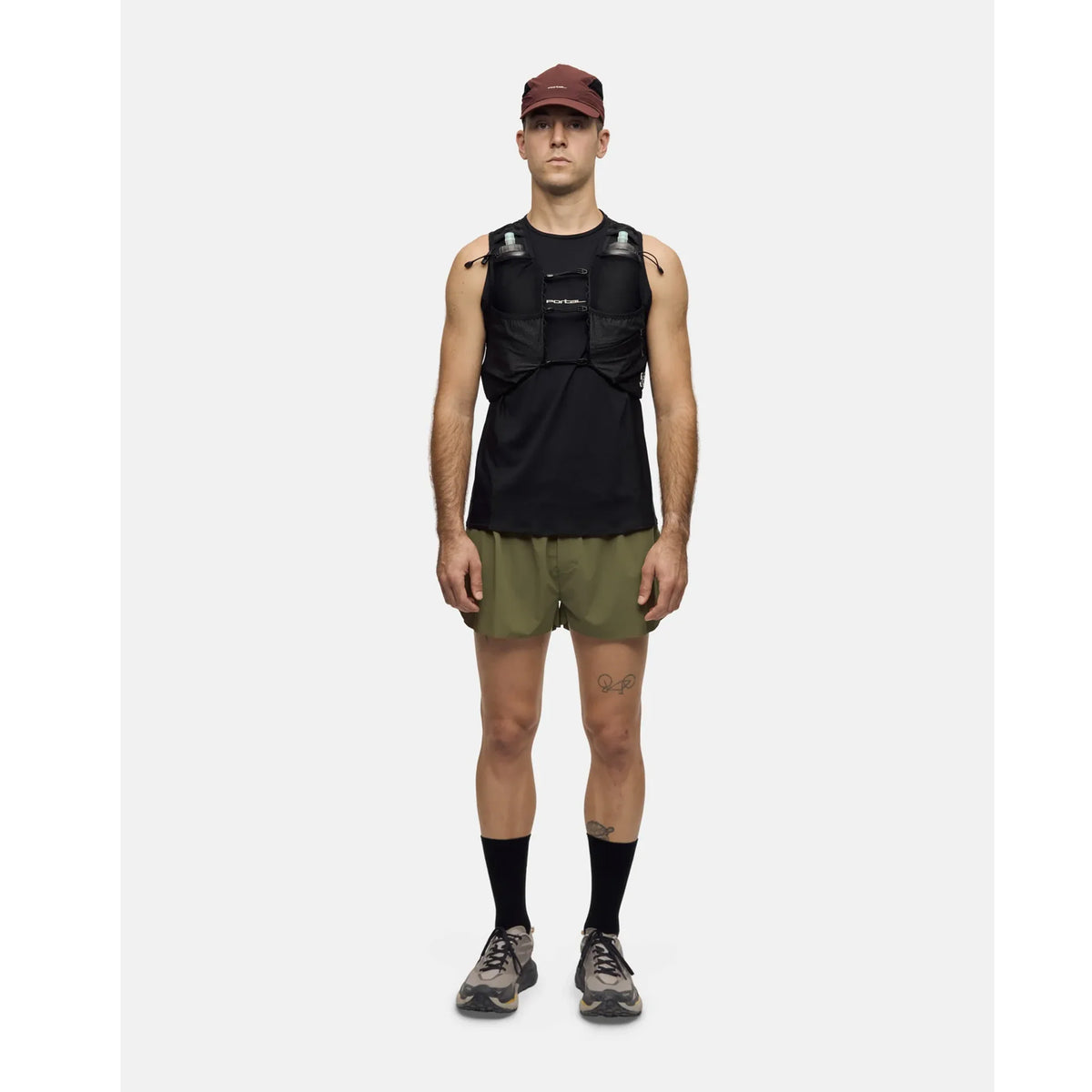 A man stands facing forward against a plain white background, wearing a maroon cap, black tank top layered with the Portal Passage Cargo Vest in Black Beauty, olive green shorts, black socks, and running shoes.